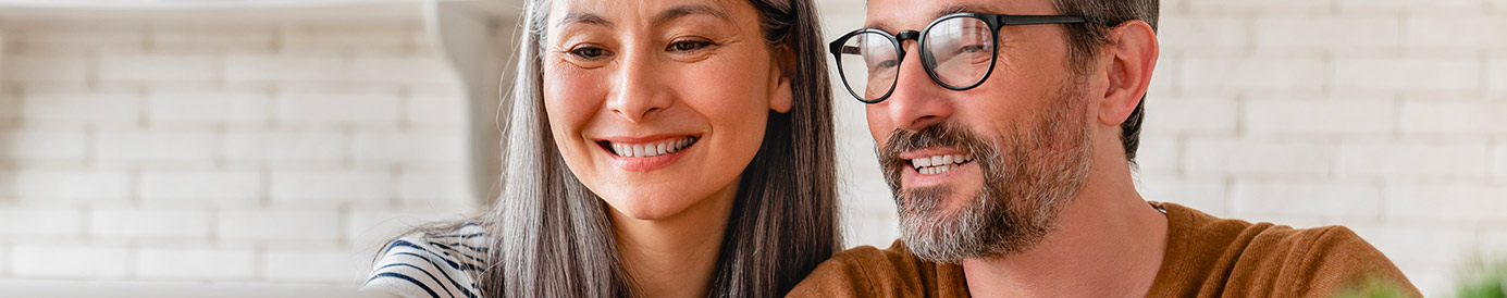 A couple sits at their kitchen table while looking at a laptop together. The man is holding a credit card while the woman points at the laptop screen.
