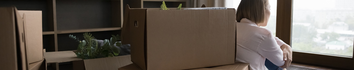 Stacks of cardboard moving boxes sit on the floor as a woman sits next to them staring pensively at a set of windows.