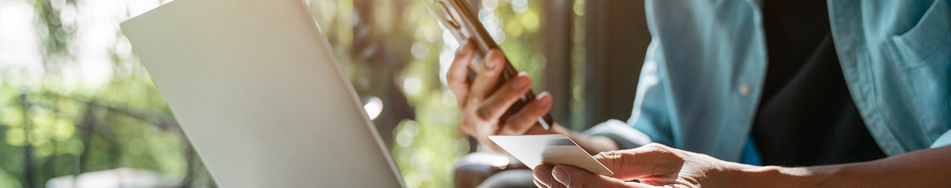A man in a coffee shop sits at a table while typing his card information into his phone. He has his phone in one hand and a credit card in the other.
