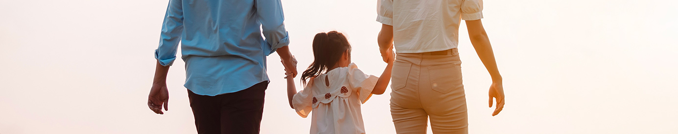 A couple and their daughter are walking in a field next to a river. The dad is on the left and the mom is on the right while they both hold the girl’s hand.