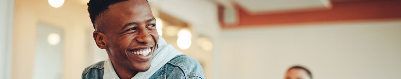 College students in a classroom, a male student at the front can be seen talking and smiling, while students behind him are blurred out.