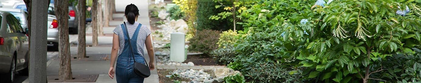 A woman wearing overalls over a striped shirt walks down a sidewalk. She is carrying a black crossover purse. There are parked cards and a row of trees to her left and townhomes to her right.