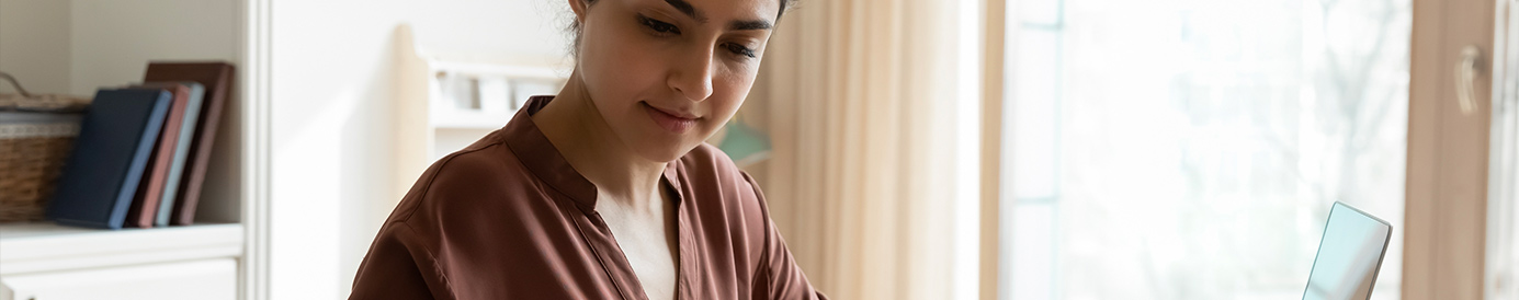A woman seated at a table is using a calculator and a laptop. She has a few papers in front of her and is calculating some numbers.