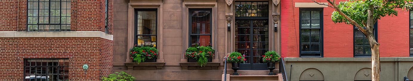 Three rowhouses in a city. The center townhouse has a brown stone exterior and a wrought iron front door with plants on the front steps.