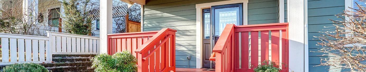 The front of a small green and brown house. The front entrance has a small, red front porch and a stone path with some plants around it.