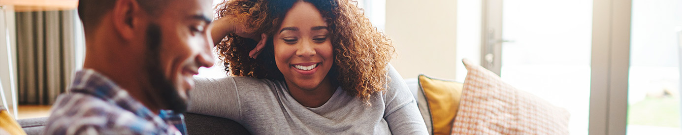 A couple sits on the couch together. The man is showing the woman something on his phone.