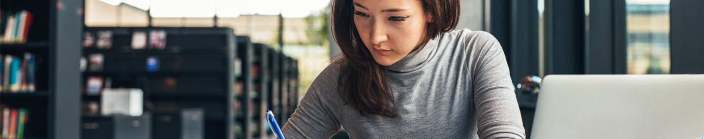 A female with long dark hair sits at a table in a library writing in a notebook. Other books and a laptop are on the table.