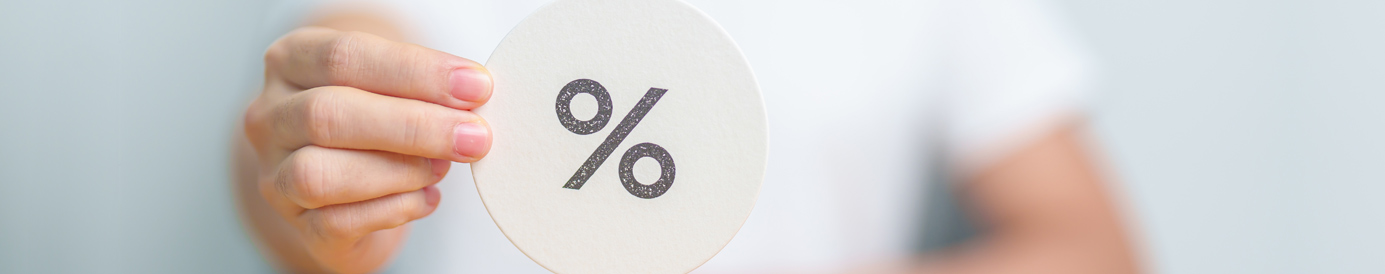 A woman in a white shirt holds a round sign with a percentage symbol in front of her. The sign is in focus and she is blurred.