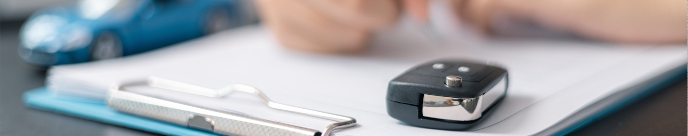 A car key fob sits on a document in a clipboard that is being signed by a person at a desk. A blue toy car sits next to it.