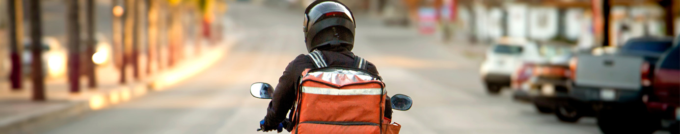 A delivery driver wearing a black helmet on a scooter rides down a street with an insulated bag resting on the back.