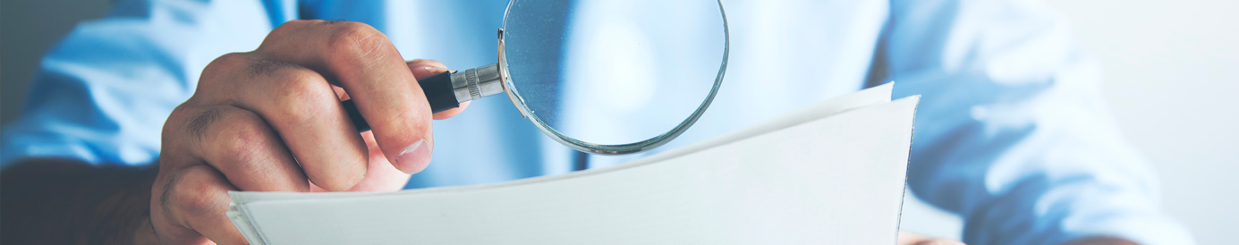 A man sits at a table with a calculator. He uses a magnifying glass in one hand to study pages of documents in the other hand.