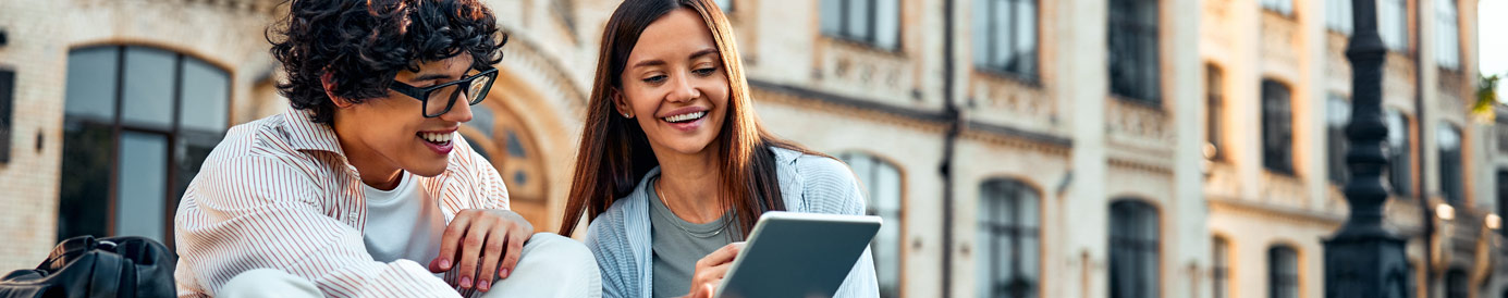 Two college students &mdash; one male and one female, sit on a campus lawn. They're smiling, laughing and looking at a tablet.