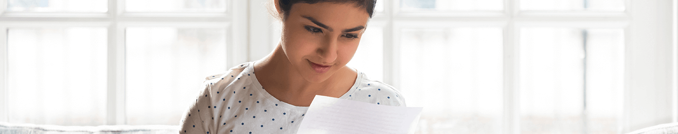 A young woman with dark hair and a white shirt looks down at a piece of paper in her hand, examining information on it.