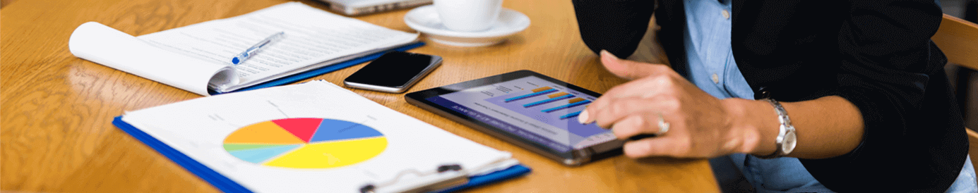 A woman sits at a desk while reading graphs and reports on an iPad. There are several papers and notebooks on the desk.