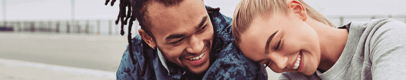 A man and woman sit on a sidewalk smiling at each other. The woman is resting her head on the man’s shoulder.