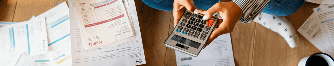 A couple sits on the floor with financial papers and invoices spread out on the floor around them. The man is holding up an invoice and the woman is typing into a calculator.