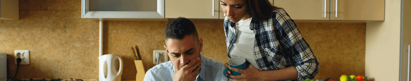 A man sits at a kitchen table looking at a paper and looks distressed. A woman stands over him also looking at the paper.