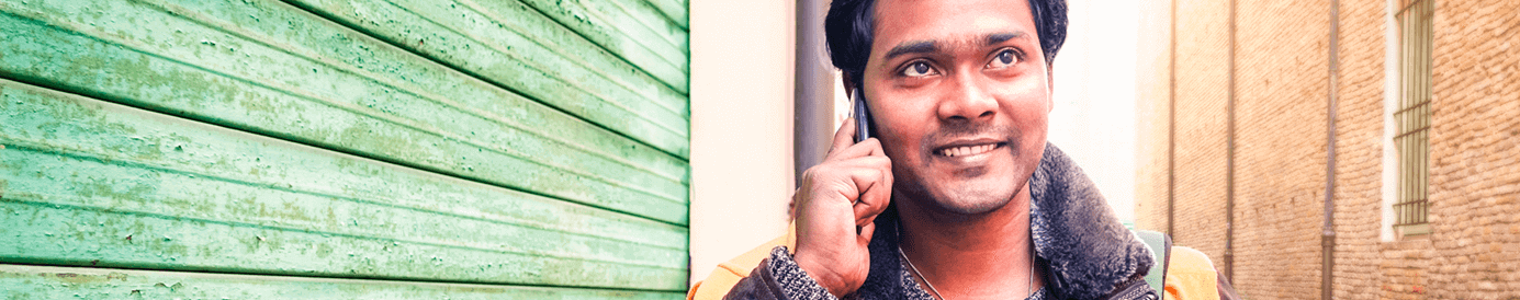 A man is walking down a city street while talking on the phone. He is wearing a coat and sweater and has a backpack on.