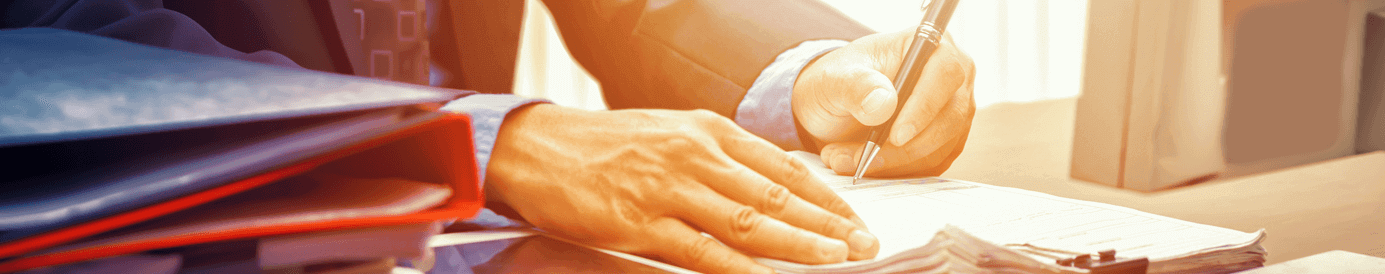 A man in a suit sits at a desk with paperwork and a tablet in front of him. He writes on the top page of a stack of papers.