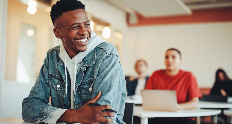 College students in a classroom, a male student at the front can be seen talking and smiling, while students behind him are blurred out.