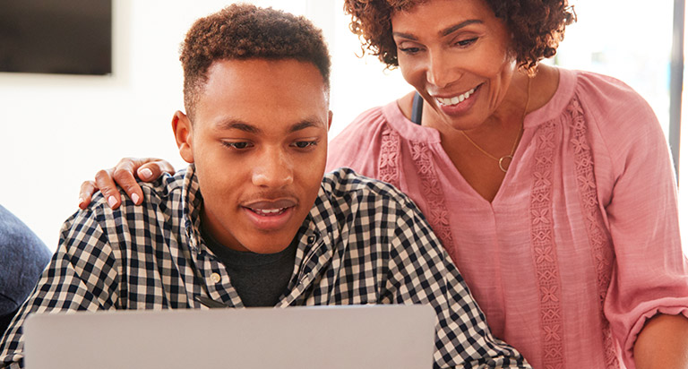 A man and woman are looking at a laptop together. The man is sitting down at the counter, while the woman stands next to him.