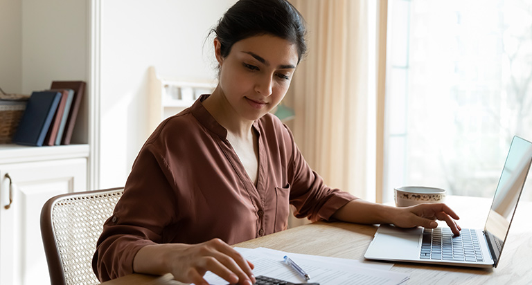 A woman seated at a table is using a calculator and a laptop. She has a few papers in front of her and is calculating some numbers.