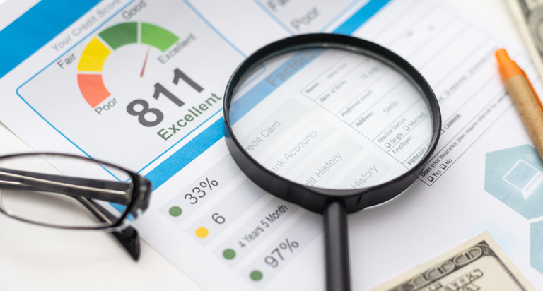 A magnifying glass rests on a surface with documents related to credit. A pen, glasses and a 100 dollar bill lay beside it.