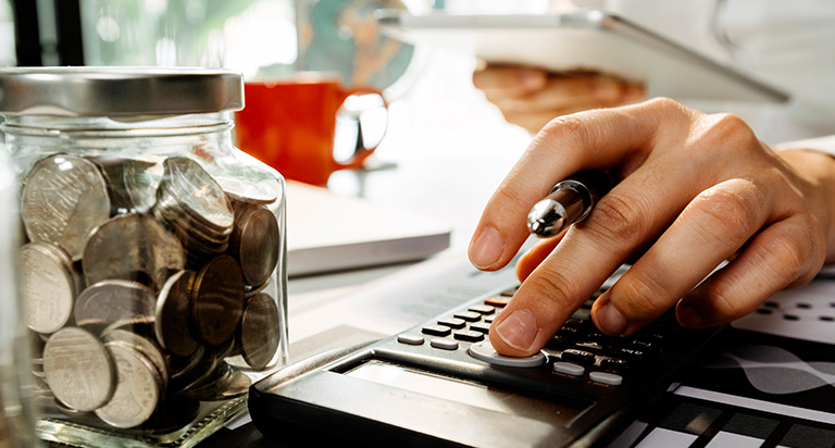 A person is reading from their iPad while typing into a calculator. There is a coffee mug and some piggy banks next to the calculator on the desk.