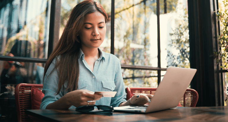 A woman sitting at a table inside a shop is reading her credit card information with a laptop in front of her.