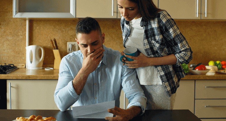 A man sits at a kitchen table looking at a paper and looks distressed. A woman stands over him also looking at the paper.